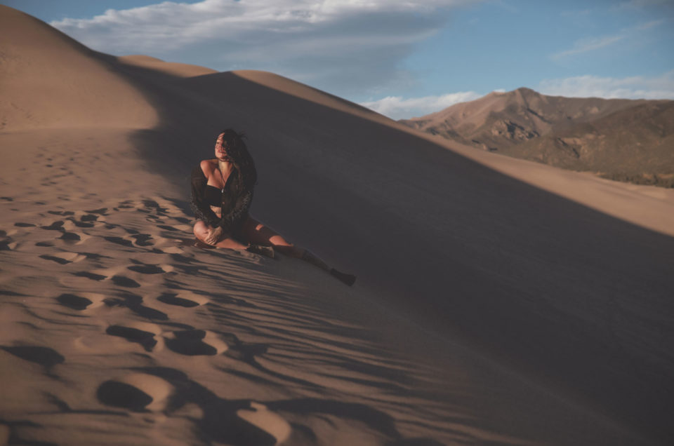 Dark-haired-woman-dressed-in-black-sitting-on-the-edge-of-a-large-sand-dune-looking-into-the-sun