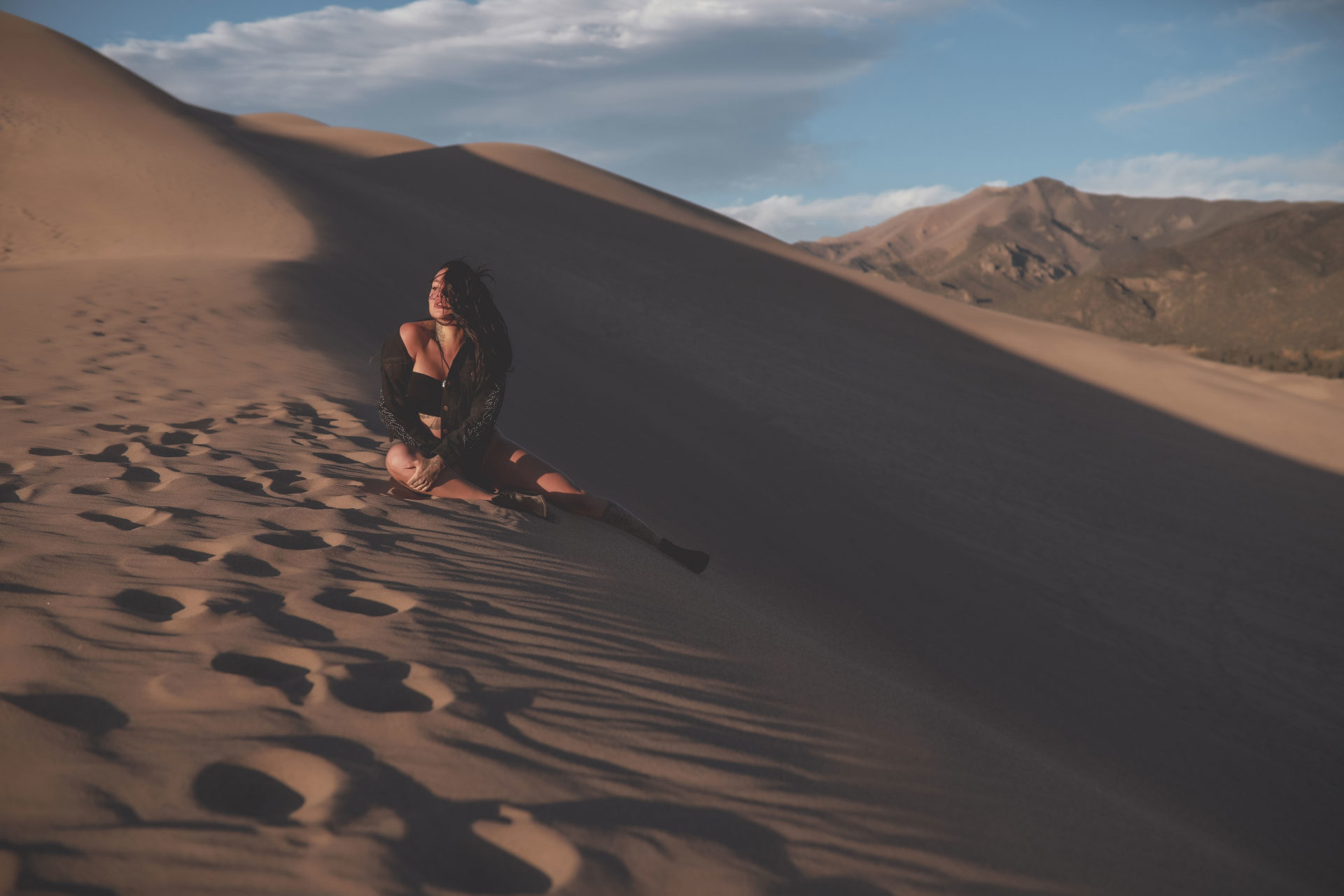 Dark-haired-woman-dressed-in-black-sitting-on-the-edge-of-a-large-sand-dune-looking-into-the-sun