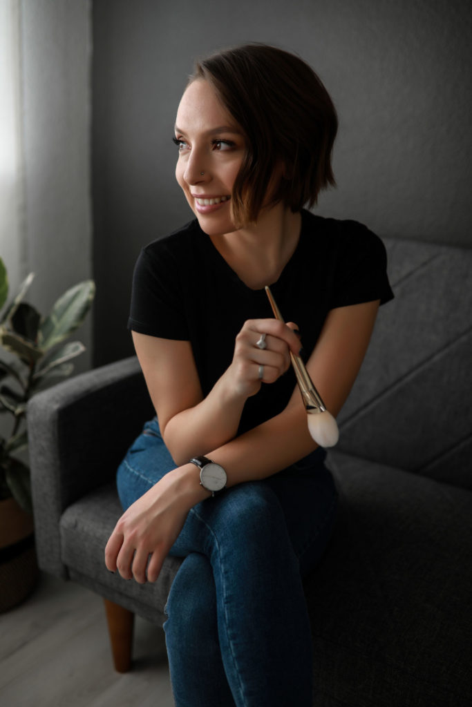 Dark-short-haired-woman-holding-a-makeup-brush-sitting-on-a-couch-smiling-looking-to-the-side