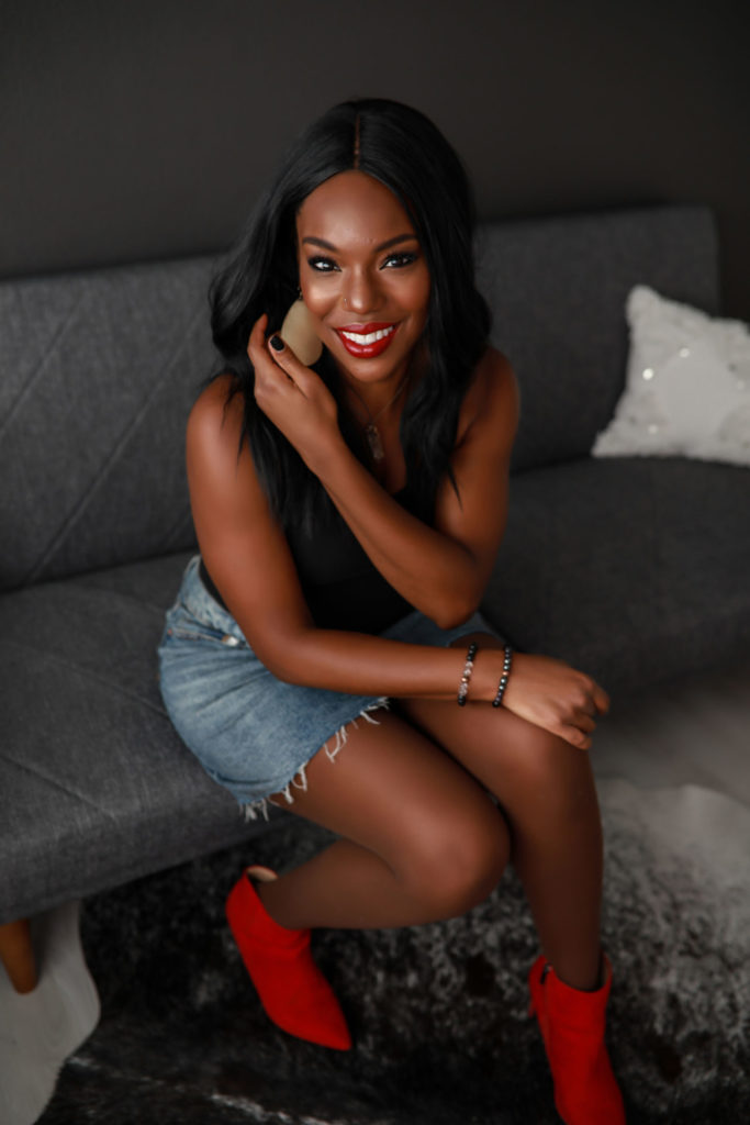 Woman-with-long-black-hair-in-a-denim-skird-black-top-and-red-boots-sitting-on-grey-couch-tucking-her-hair-behind-ear