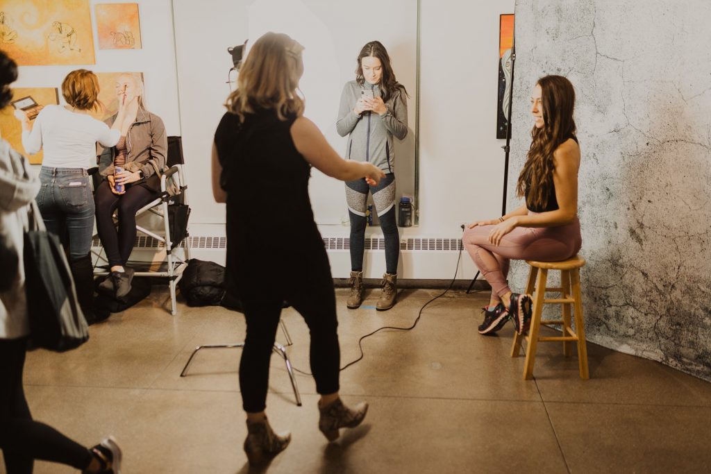 Woman photographer getting ready to take an image of a brown haired woman in a yoga outfit sitting on a wood stool. 