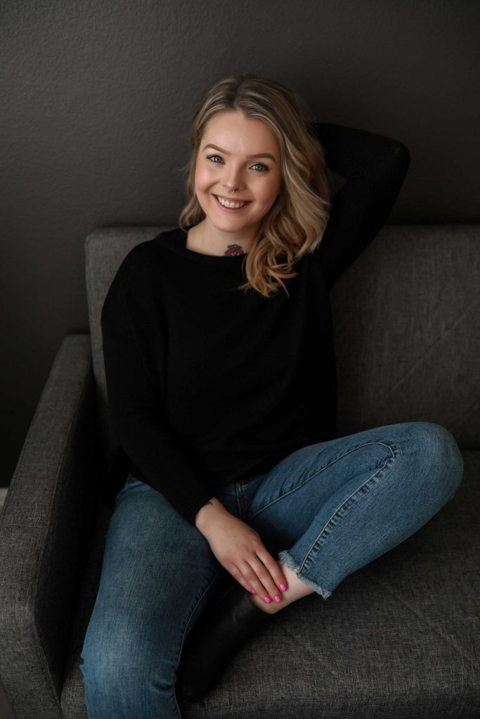 Professional-headshot-of-blonde-woman-weaing-black-sweater-and-jeans-posing-on-a-grey-couch