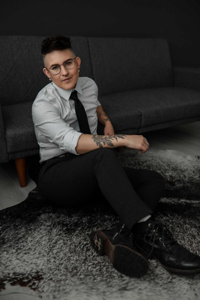 Professional-heahshot-of-a-nonbinary-wearing-a-button-up-shirt-and-dress-pants-sitting-on-the-floor-leaning-against-a-grey-couch