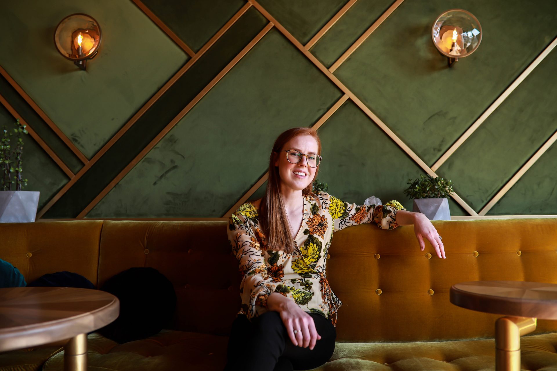 Colorado-headshot-of-woman-wearing-floral-blouse-sitting-on-a-gold-velvet-couch-with-a-green-velvet-wall-with-gold-accents