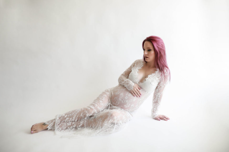 Pregnate-woman-wearing-long-white-lace-dress-posing-against-white-wall-for-her-denver-surrogacy-photoshoot