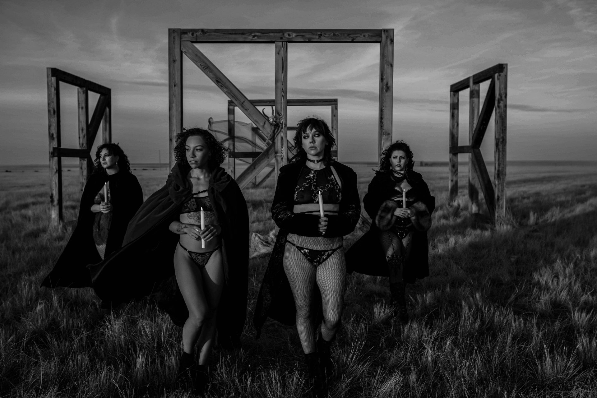 Witchy-women-dressed-in-lingerie-and-cloaks-posing-in-front-of-wooden-structures-in-the-middle-of-a-field