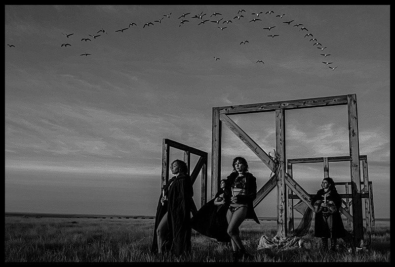 Witchy-women-dressed-in-lingerie-and-cloaks-posing-in-front-of-wooden-structures-in-the-middle-of-a-field