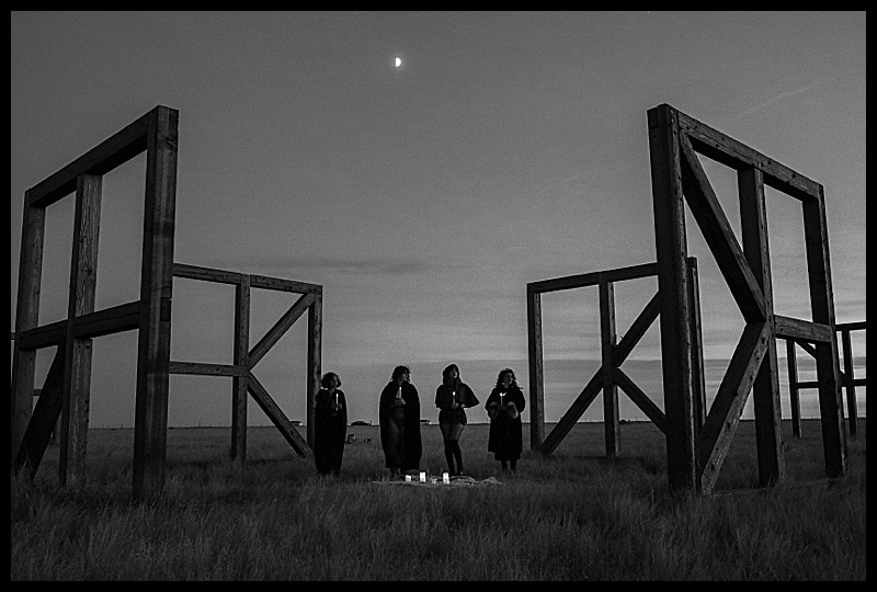 Witchy-women-dressed-in-lingerie-and-cloaks-posing-in-front-of-wooden-structures-in-the-middle-of-a-field