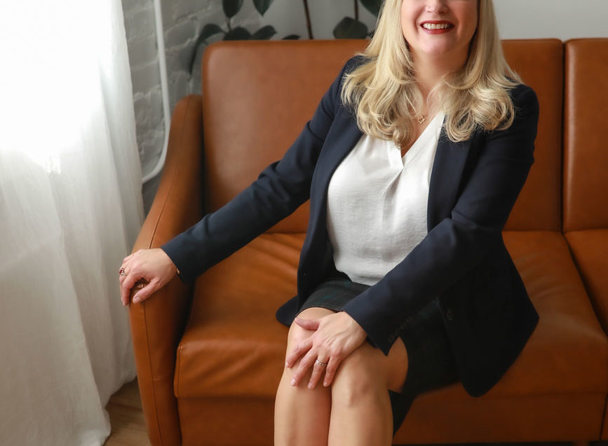 A professional woman in a business suit smiling while seated on a brown sofa in a room with a plant and window, perfect for Denver lifestyle headshots.