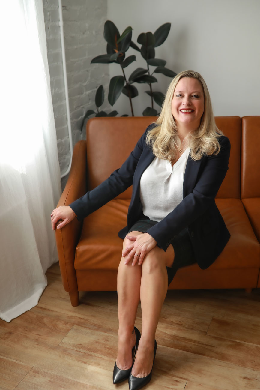 A professional woman in a business suit smiling while seated on a brown sofa in a room with a plant and window, perfect for Denver lifestyle headshots.