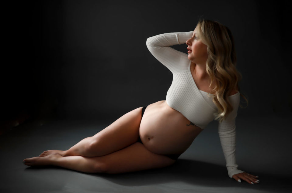 Pregnate-blonde-woman-wearing-white-crop-top-and-black-bottoms-posing-on-floor-of-dark-backdrop