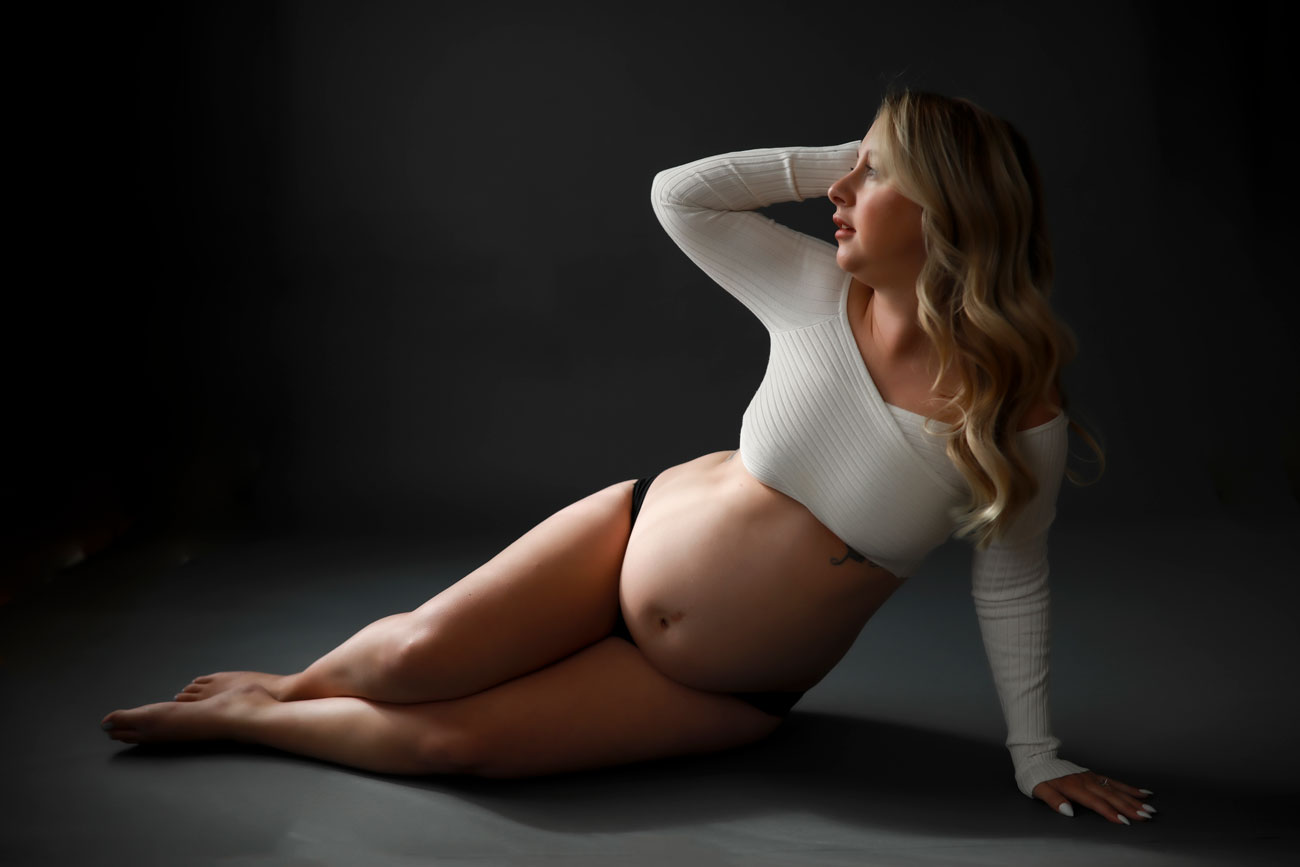 Pregnate-blonde-woman-wearing-white-crop-top-and-black-bottoms-posing-on-floor-of-dark-backdrop