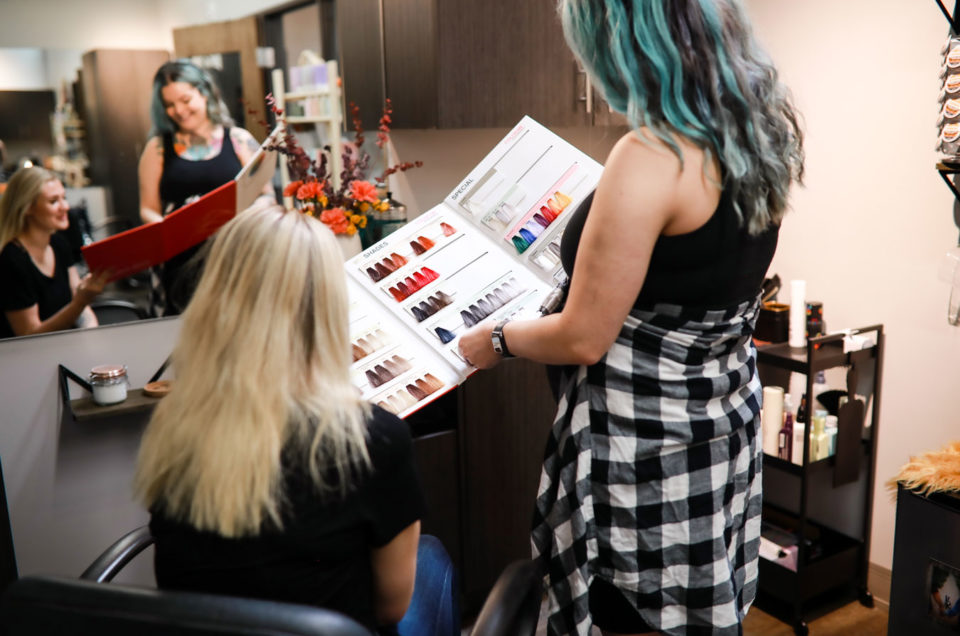 A Denver hairstylist holding a hair color sample chart shows options to a seated client in a salon.