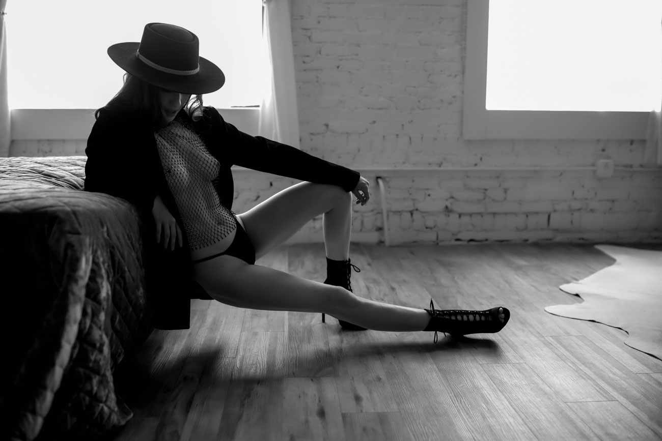 Woman in heels, mesh top with blazer posing on edge of bed.