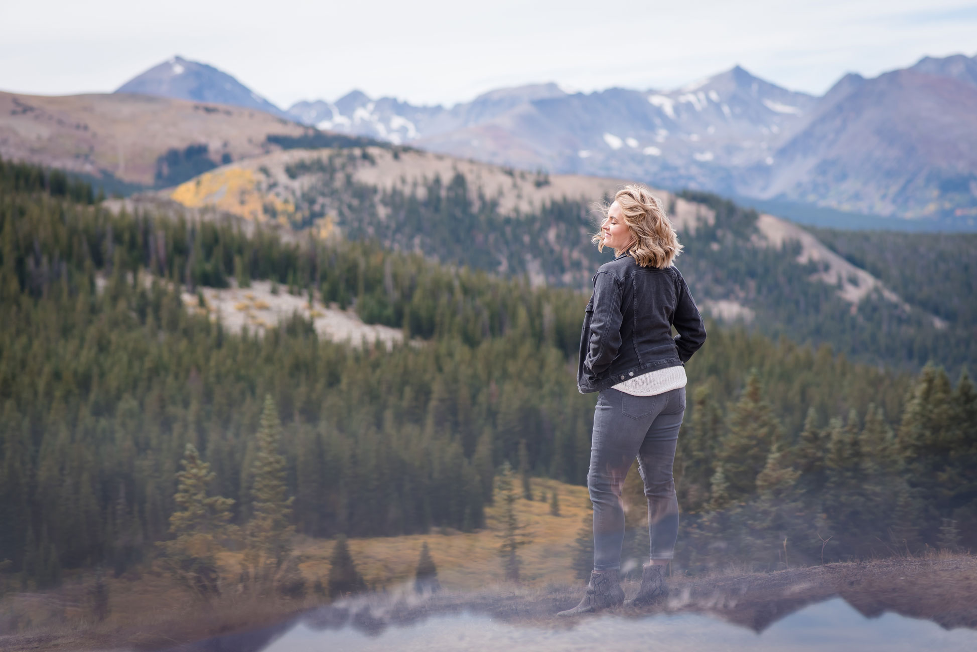 Woman in all black denim with hands in pockets overlooking piney forest.