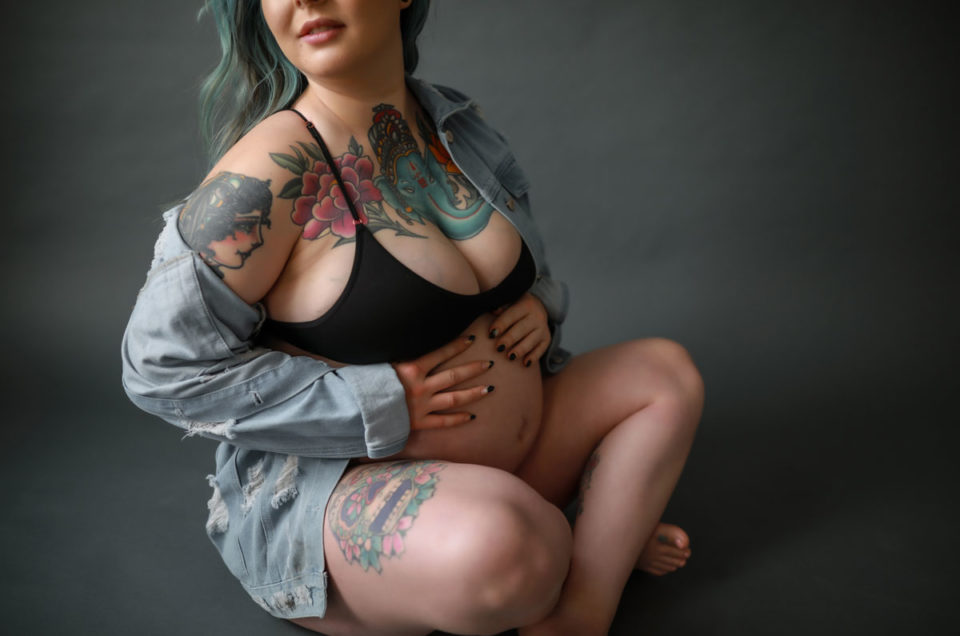 Pregnant tattooed woman sitting on floor wearing denim jacket.