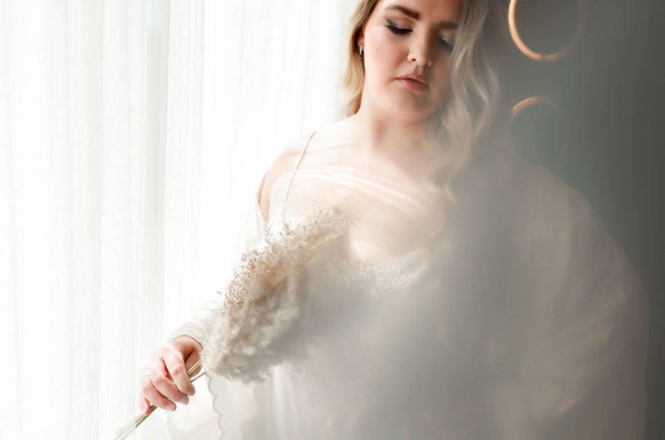 Boudoir image of woman holding pampas grass wearing white robe.