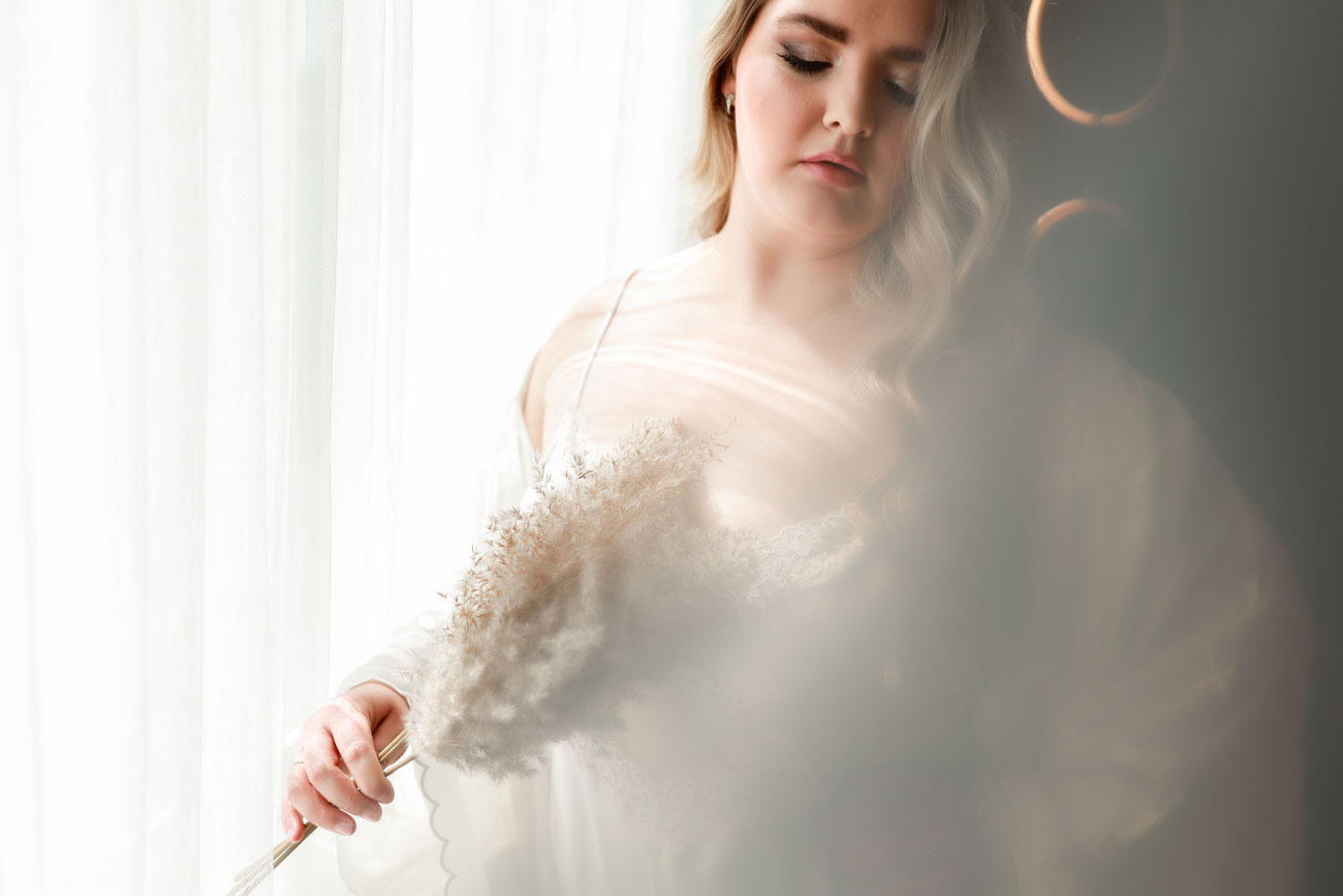 Boudoir image of woman holding pampas grass wearing white robe.