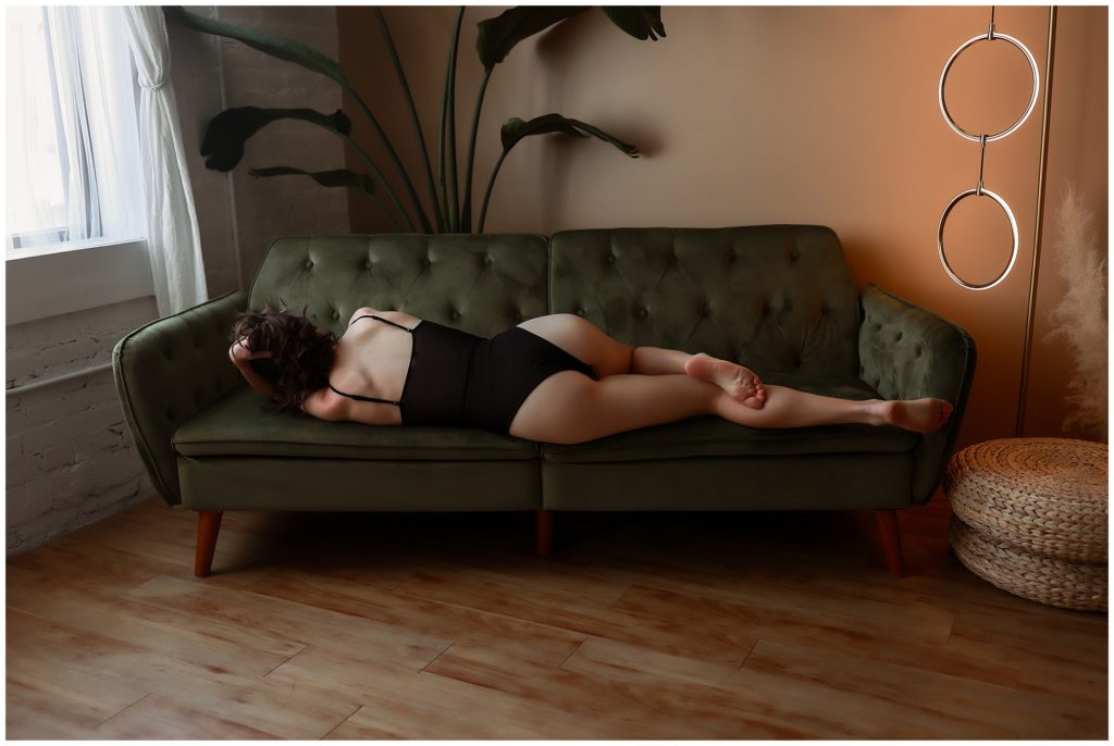 Woman in black bodysuit laying on green velvet couch.