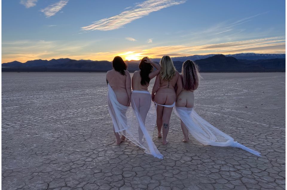 Four women standing with their backs to the camera, draped in sheer fabrics, watching the sunset in a desert landscape during The Boudoir Summit.