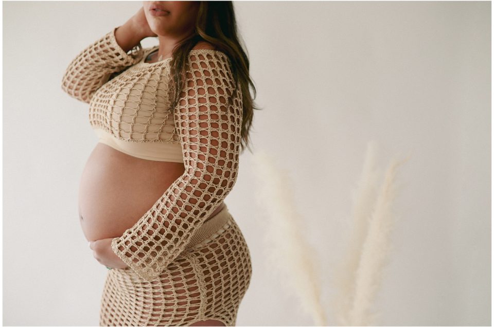 A pregnant individual in a patterned outfit cradling their belly, standing against a neutral backdrop, captures the best time to take maternity photos.