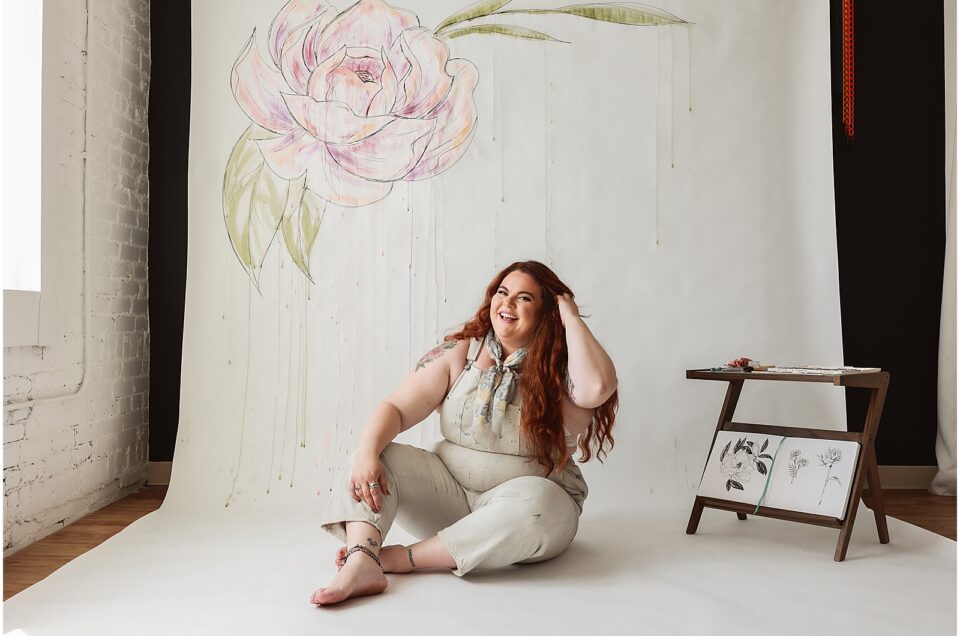 Woman sitting on the floor during a branding photoshoot, smiling, with a painted flower on the wall behind her.