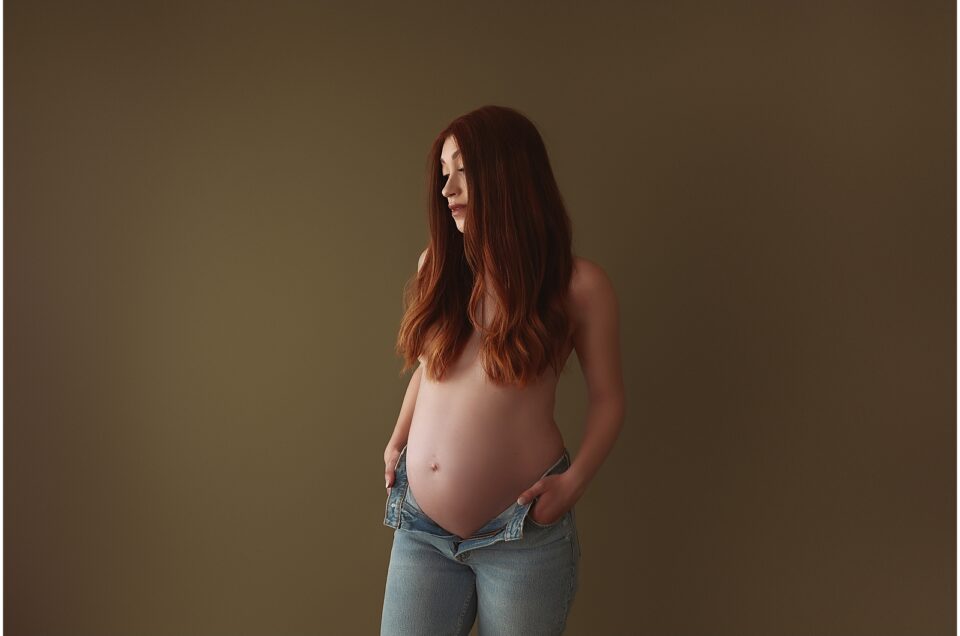 Pregnant person with long red hair participating in a maternity photoshoot, standing to the side and looking down at their belly against a brown background.