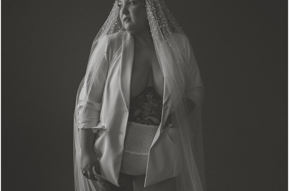 A black and white Denver Bridal Boudoir portrait of a bride in a veil and robe, revealing undergarments and a tattoo on her ribcage.