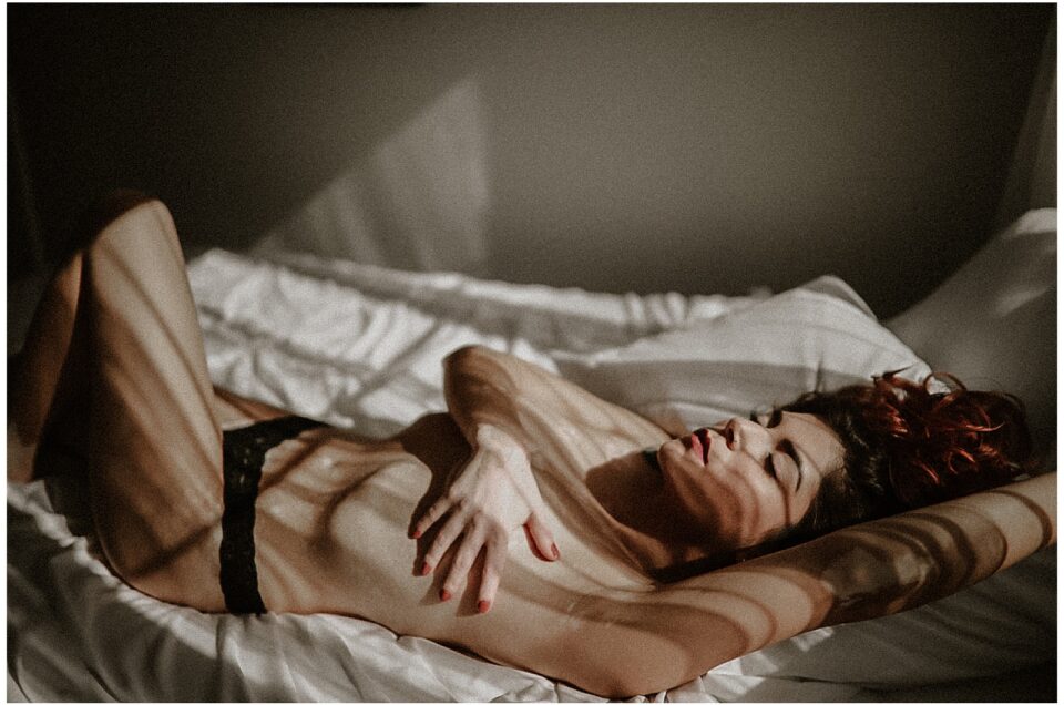 A person is lying on a bed with closed eyes, bathed in shadows and light, creating a contrast on their body, encapsulating a boudoir experience.