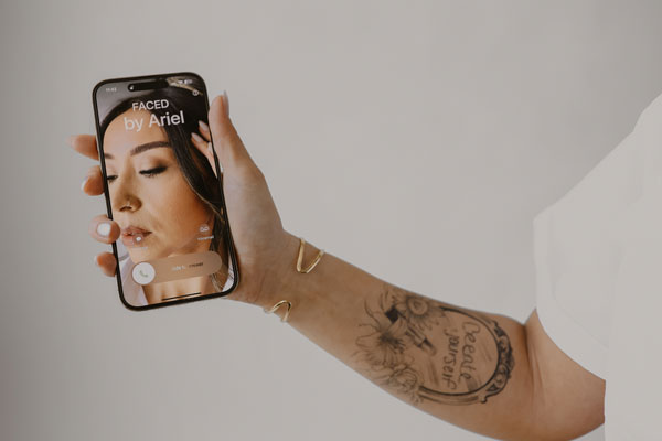 A person's tattooed arm holding up a smartphone with a Denver Makeup Artist's tutorial on the screen.