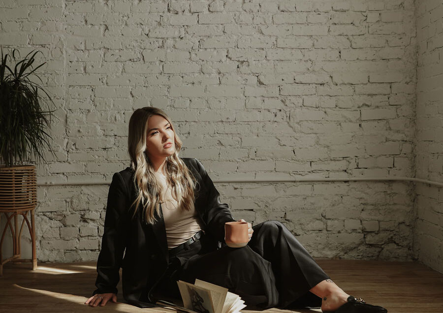 A woman in a black outfit and loafers sits on a wooden floor, leaning against a white brick wall, holding a cup and looking away from an open book.