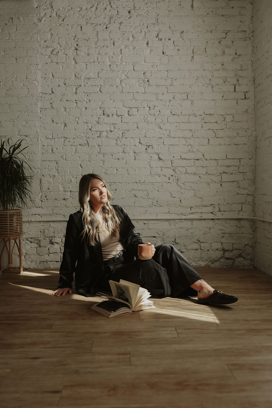 A woman in a black outfit and loafers sits on a wooden floor, leaning against a white brick wall, holding a cup and looking away from an open book.