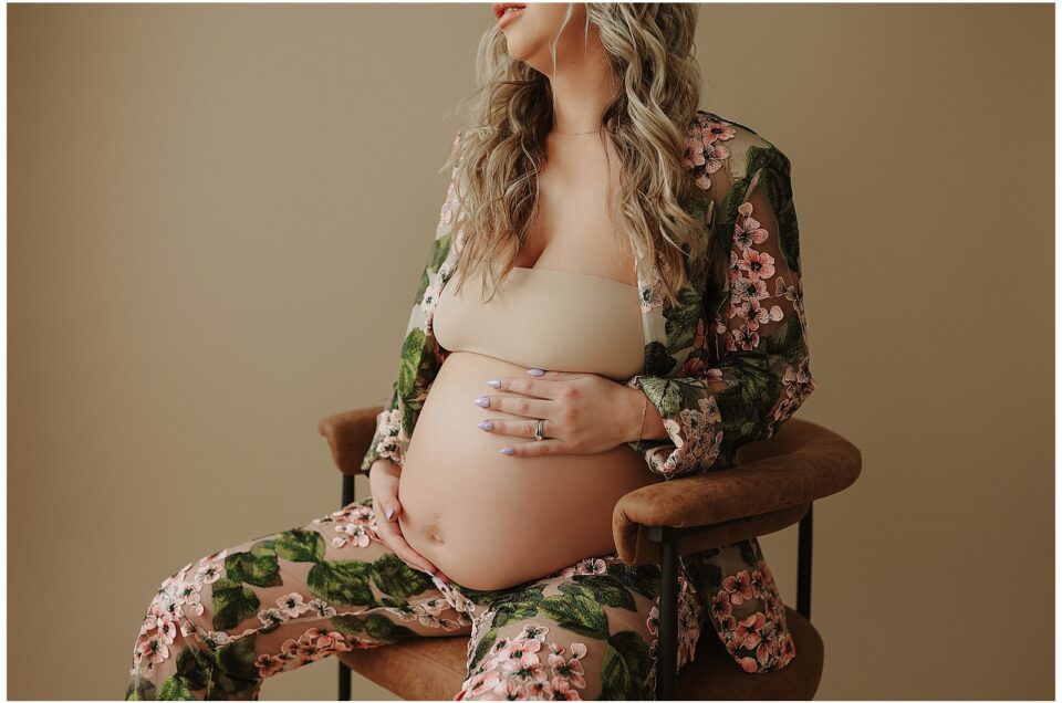 Pregnant woman in a Chelsea Football Club robe sitting on a chair, cradling her belly, with a neutral backdrop.