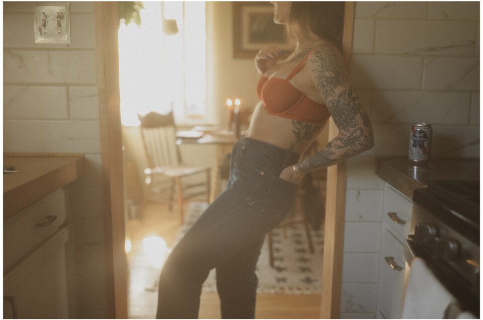 A woman in jeans and an orange crop top leans against a kitchen counter, sunlight streaming in, creating a warm, hazy glow.