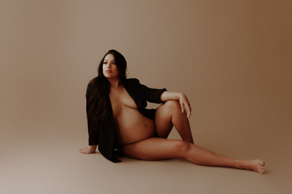 A woman with long dark hair sits on the floor, partially covered by a black blazer, in a beige-toned room.