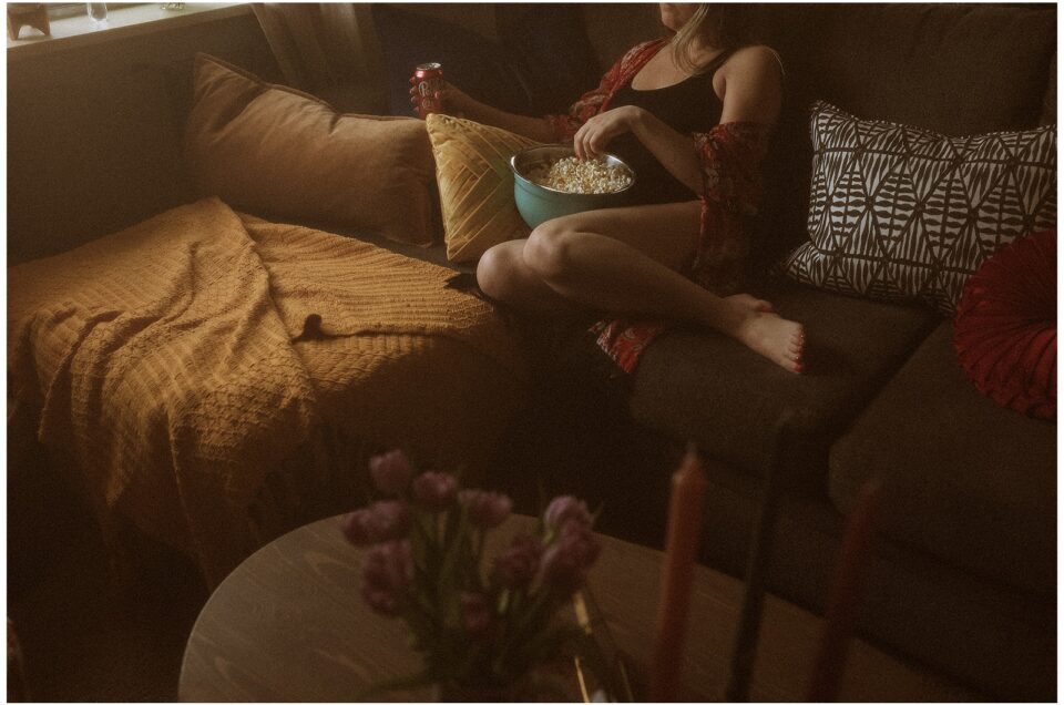 Kaitlyn relaxing on a couch with a bowl of popcorn, surrounded by pillows in a dimly lit room. There's a table with flowers in the foreground.