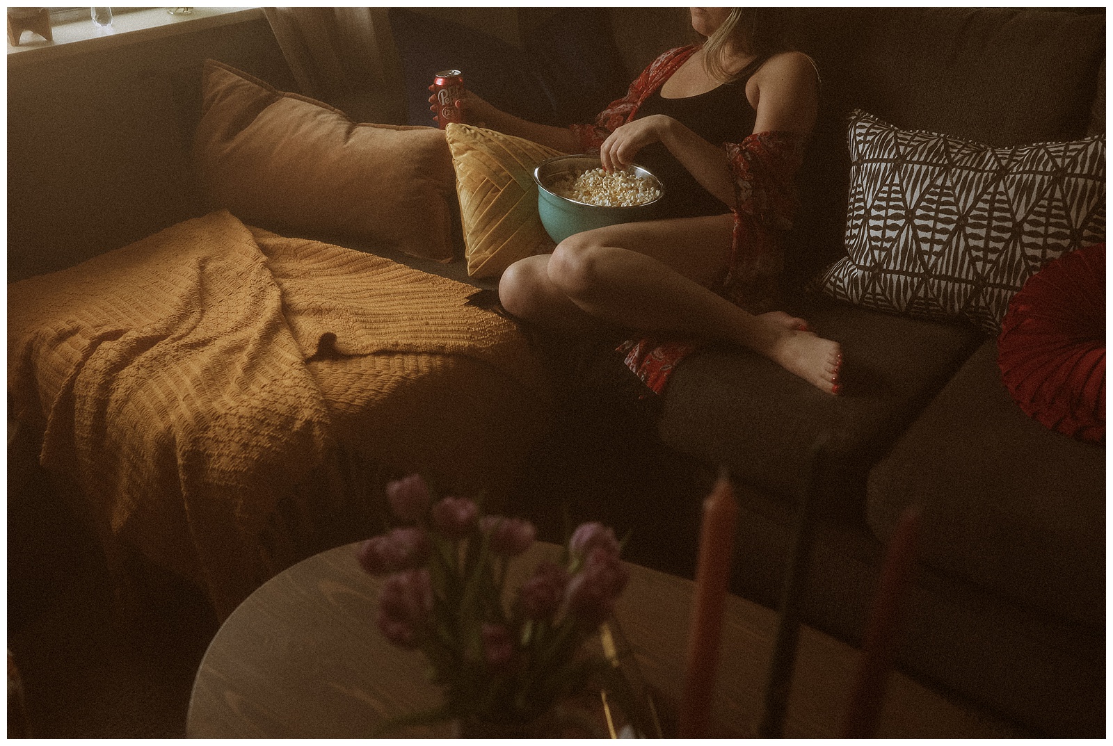 Kaitlyn relaxing on a couch with a bowl of popcorn, surrounded by pillows in a dimly lit room. There's a table with flowers in the foreground.