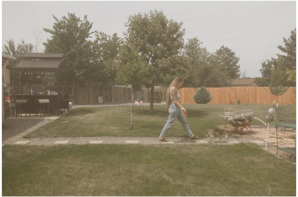 In a moment of reverie, a woman walks a small dog across a grassy backyard. An intimate scene unfolds with a shed and fenced area in the background, under an overcast sky.