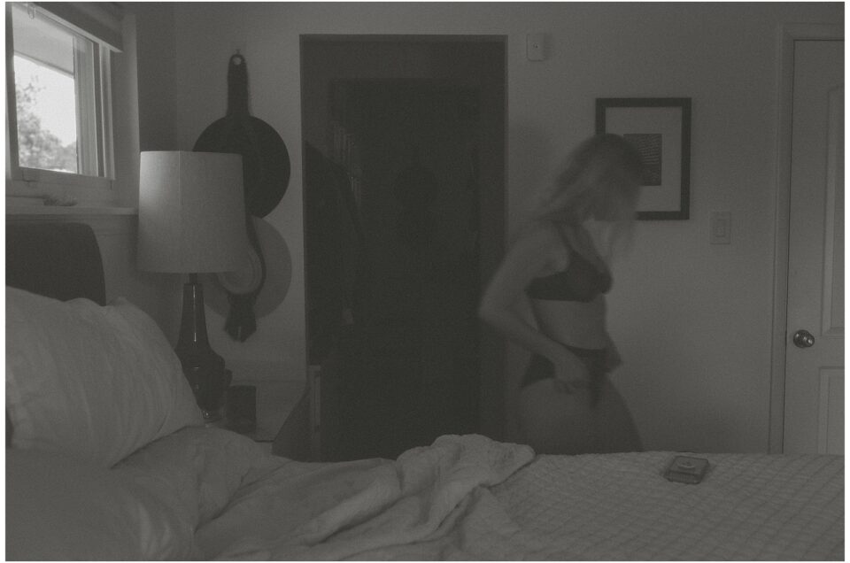 A woman in underwear stands beside an unmade bed in a dimly lit bedroom, lost in a reverie. A lamp and hats are visible on the left, while a doorway and a framed picture are on the right, capturing the essence of quiet summer mornings.