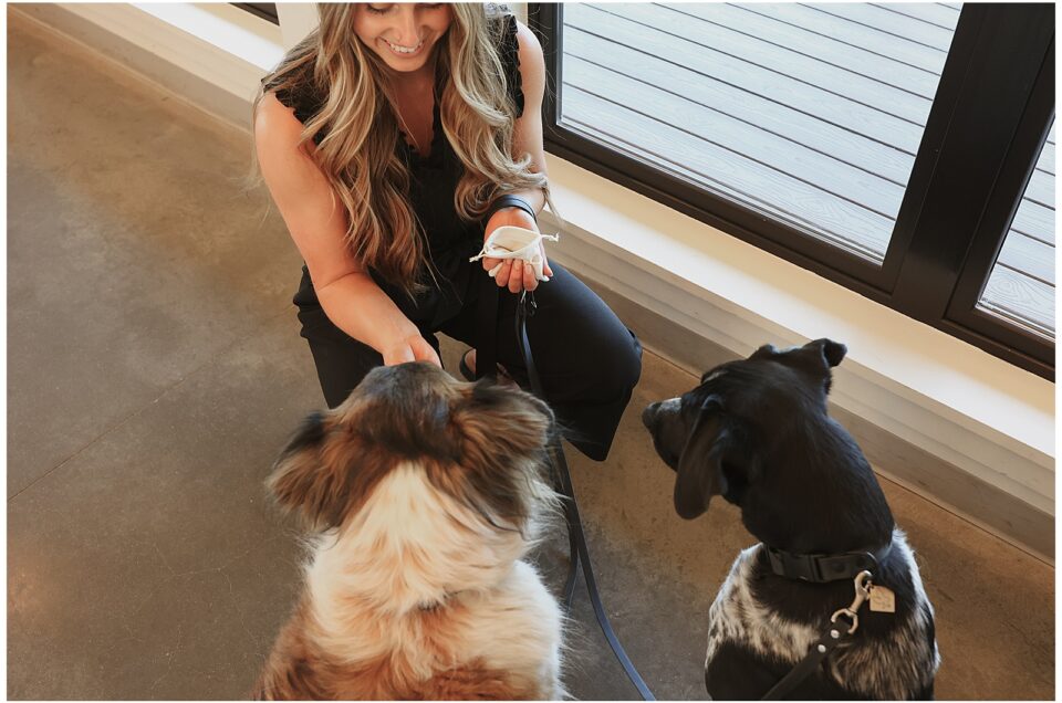 A woman kneels on the floor, smiling at two seated dogs. She holds treats in her hand as the dogs look up at her attentively.