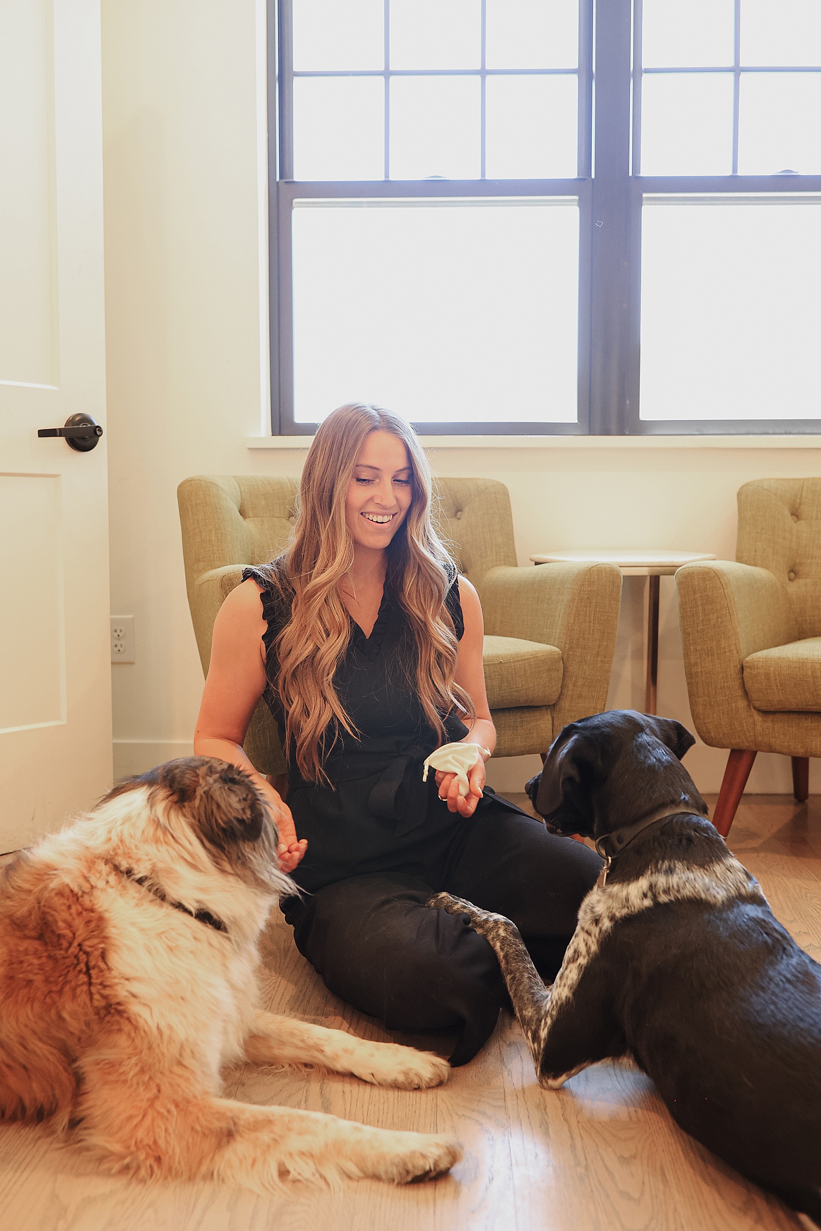 A woman kneels on the floor indoors, holding a toy, with two dogs sitting on either side of her. Two beige chairs are in the background.