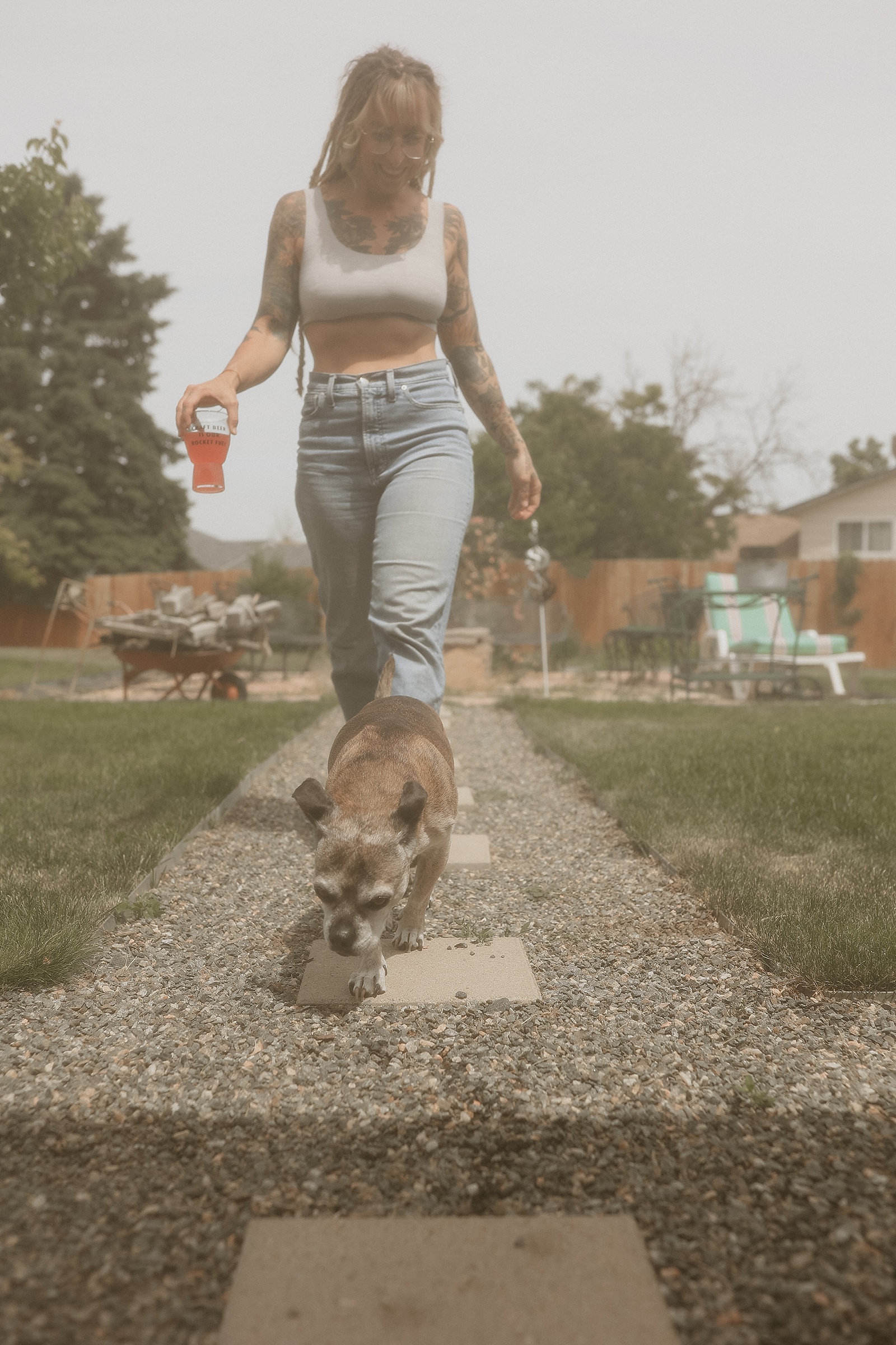 A woman with tattoos walks on a stone path in a backyard, holding a red drink, while a small dog walks ahead of her.