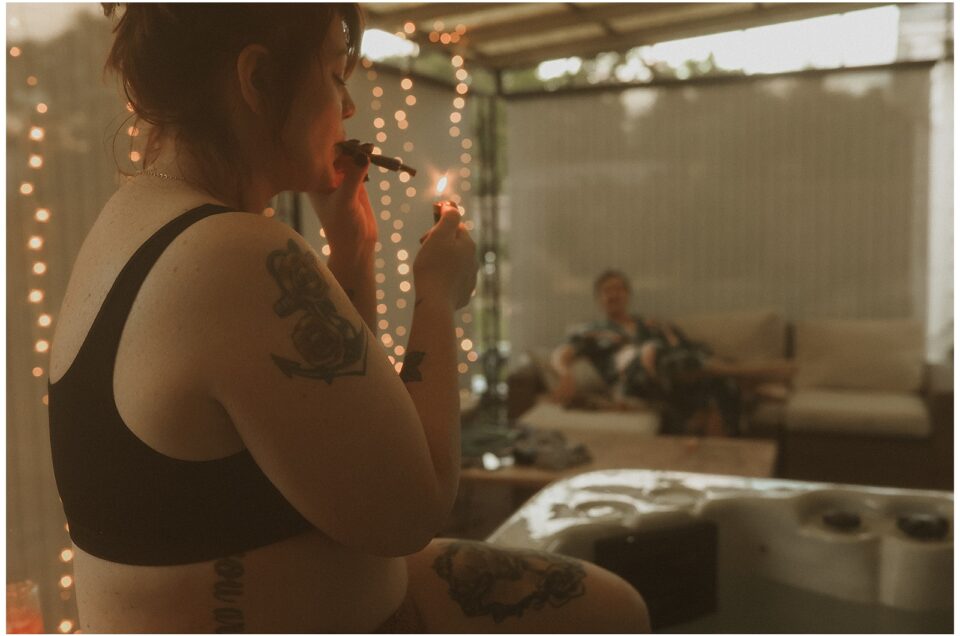 A woman in a black sports bra lights a joint in a room decorated with string lights. A man sits on a sofa in the background.