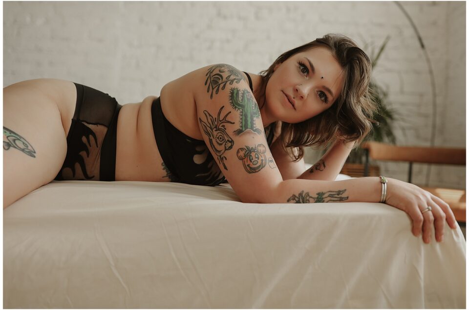 A person with tattoos lies on their side on a bed, wearing black undergarments. A brick wall and plant are visible in the background.