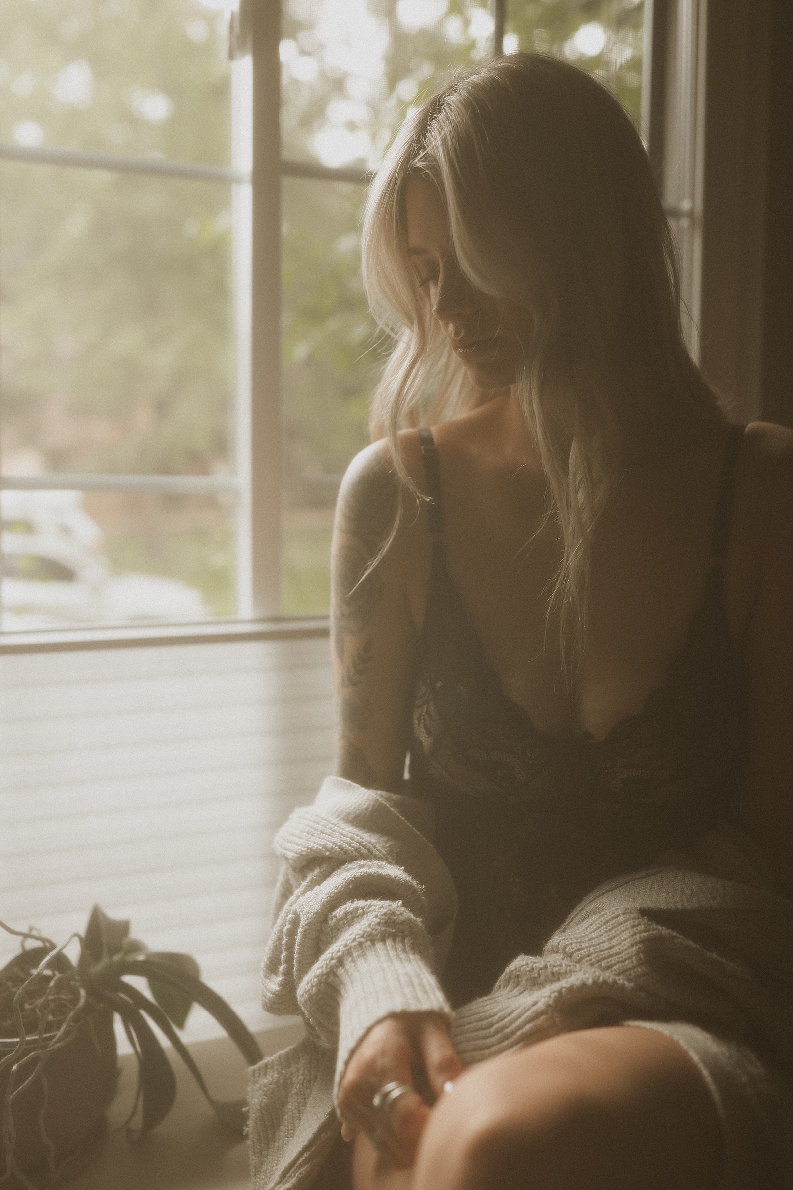 Person with long hair in a lace top sits by a window, softly lit by natural light, with a plant on the windowsill and partially covered by a sweater.