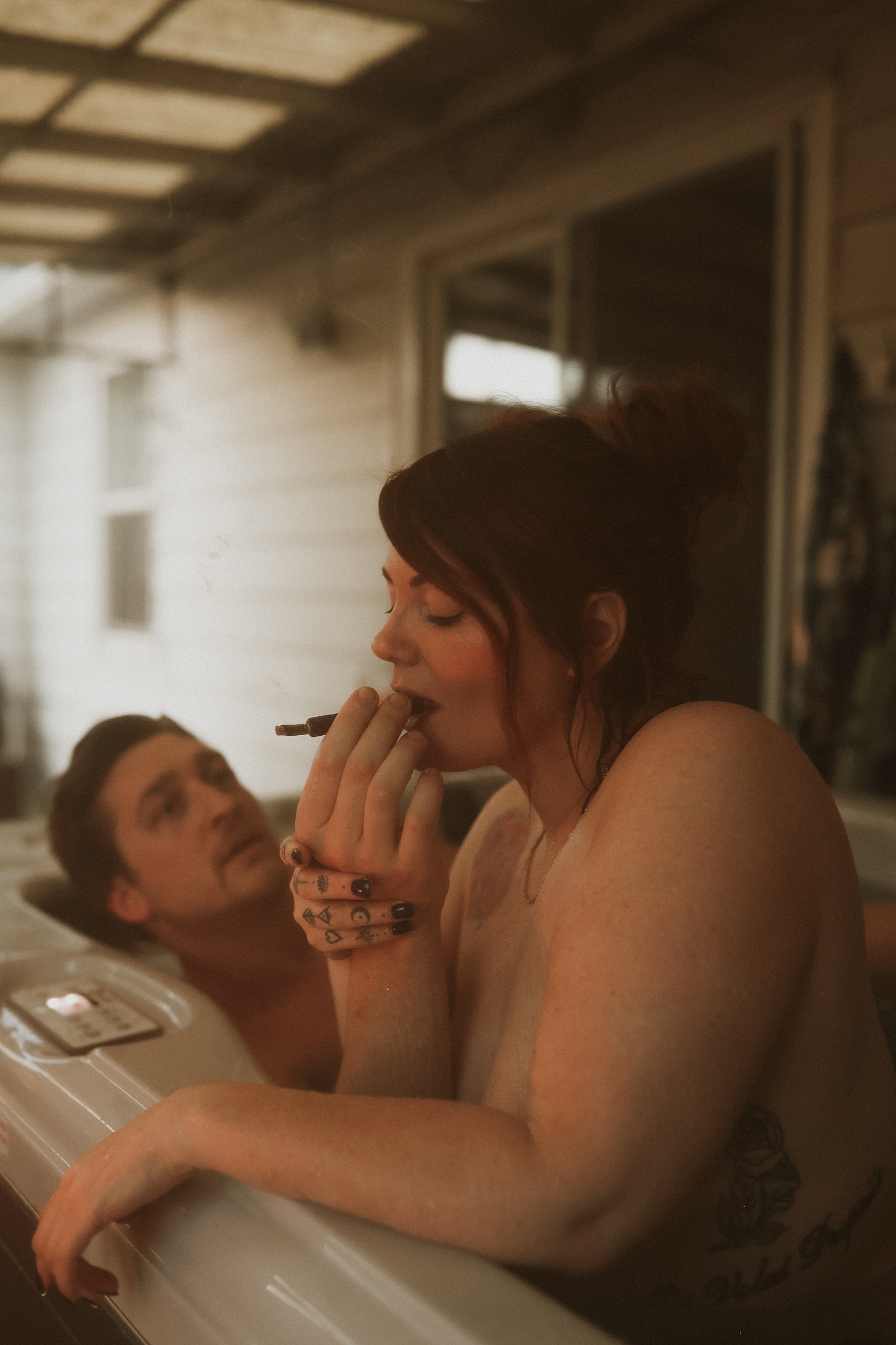 Two people in a hot tub; one person is smoking while the other looks on, indoors with a window in the background.