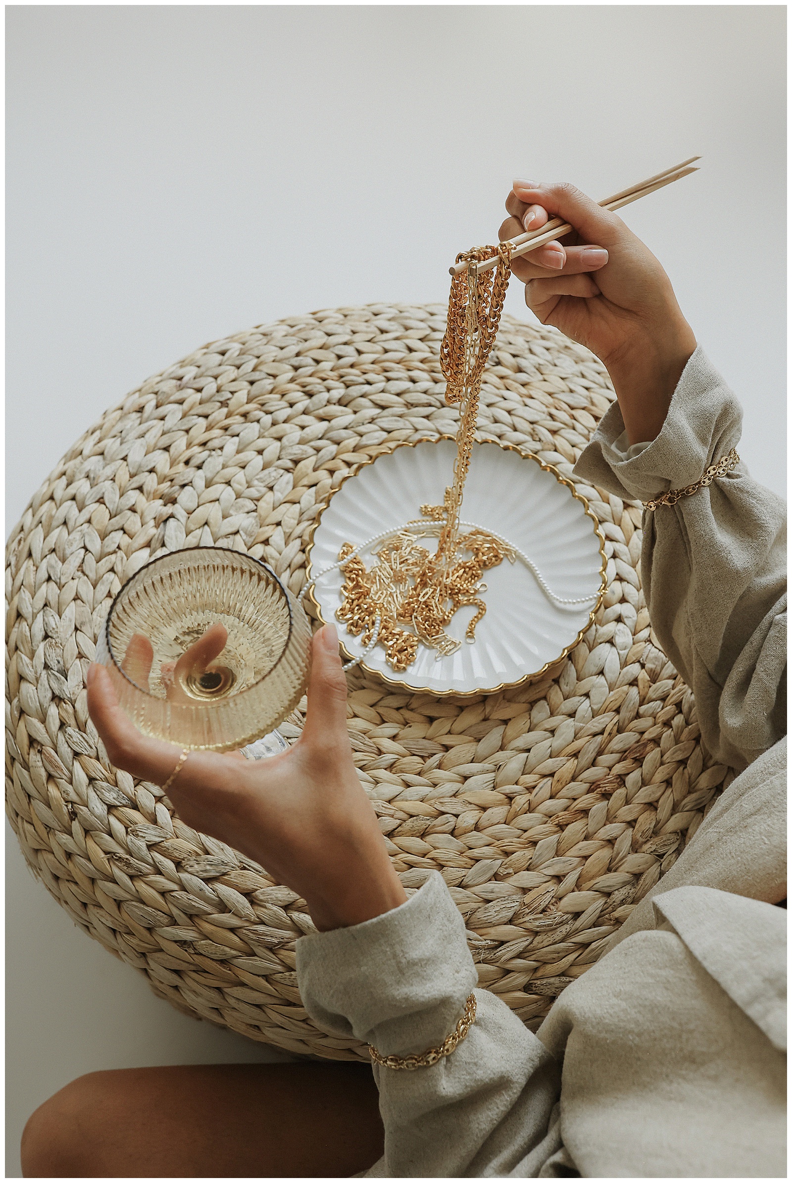 A person is holding a glass of wine in one hand and using chopsticks to hold gold necklaces over a decorative plate, all set on a woven surface.