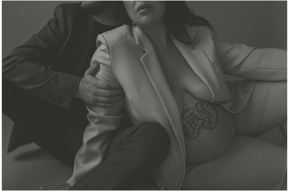 A maternity monochrome image depicts a pregnant woman with a large tattoo on her abdomen, leaning against a man, both dressed in blazers and trousers.