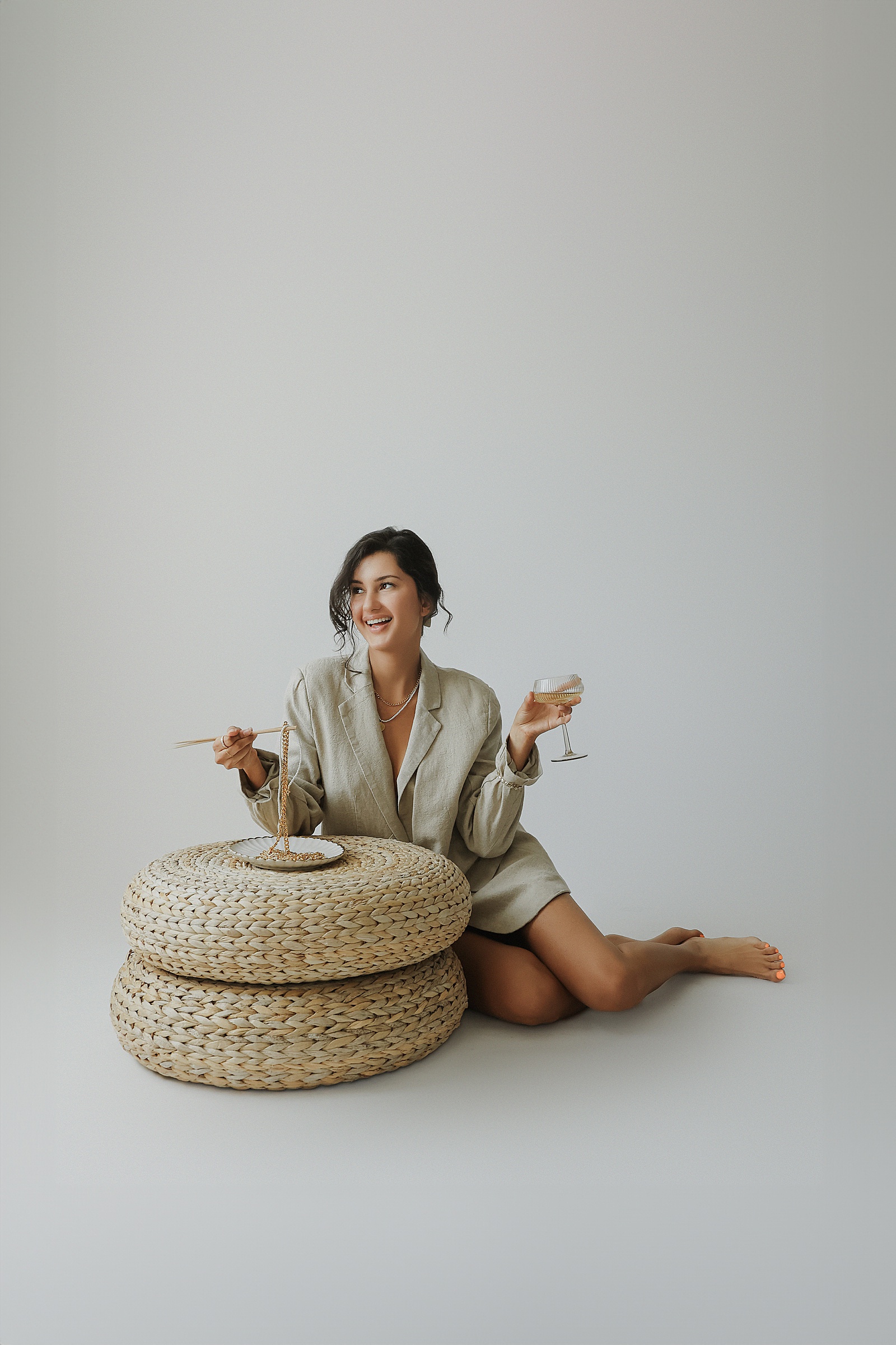 Woman sitting on the floor in a beige outfit, holding a wine glass and a bowl, with a plate on a woven ottoman.