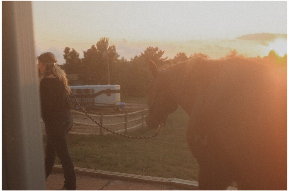 A person in reverie walks a horse on a lead rope during sunset near a fenced area, with trees and a trailer in the background.